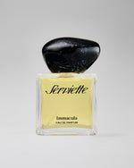 A bottle of Serviette Immacula Eau de Parfum with pale yellow liquid, a clear glass base, and a large polished black stone-like cap, centered on a light gray background.