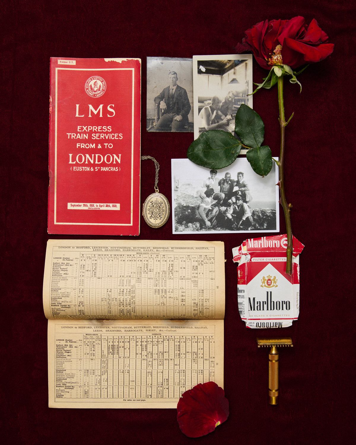A styled flat-lay of vintage memorabilia on a dark red fabric background, including an open aged timetable, a red LMS express train services booklet, black-and-white photos, a red rose, a gold pocket watch, a Marlboro cigarette pack, and a gold safety razor.
