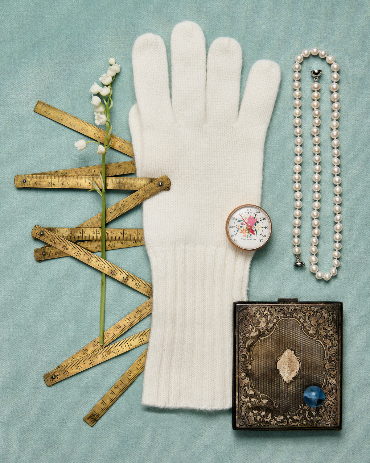 A stylized flat-lay with a white mitten, a string of pearls, a stem of lily of the valley, a yardstick that folds up and a vintage makeup compact on top of an eggshell blue background.