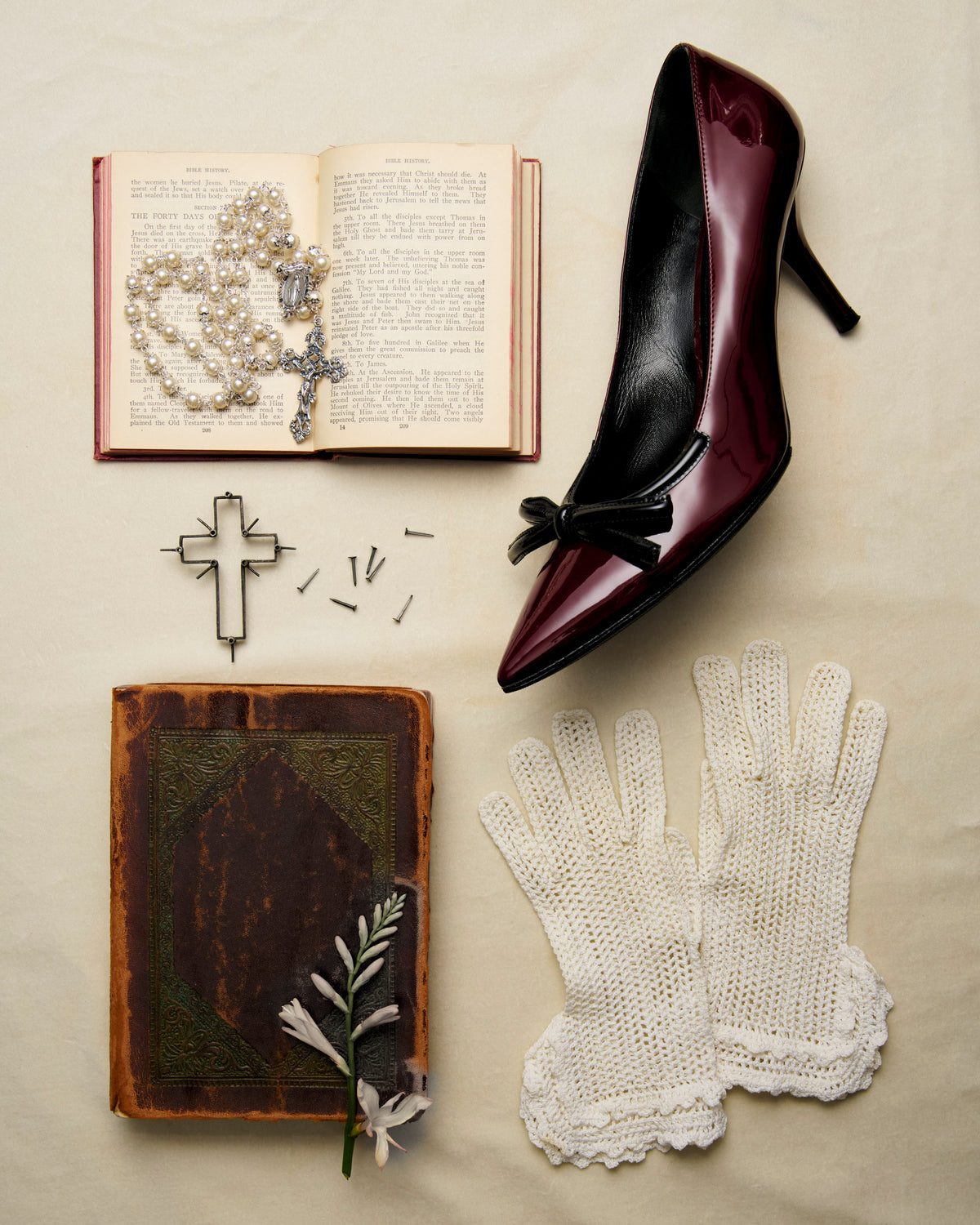 A styled flat-lay featuring a pointed-toe high heel with glossy burgundy and black panels, arranged with an open book, pearl necklace, rosary, metal cross, small nails, an old leather-bound book, and white crocheted gloves on a beige fabric background.