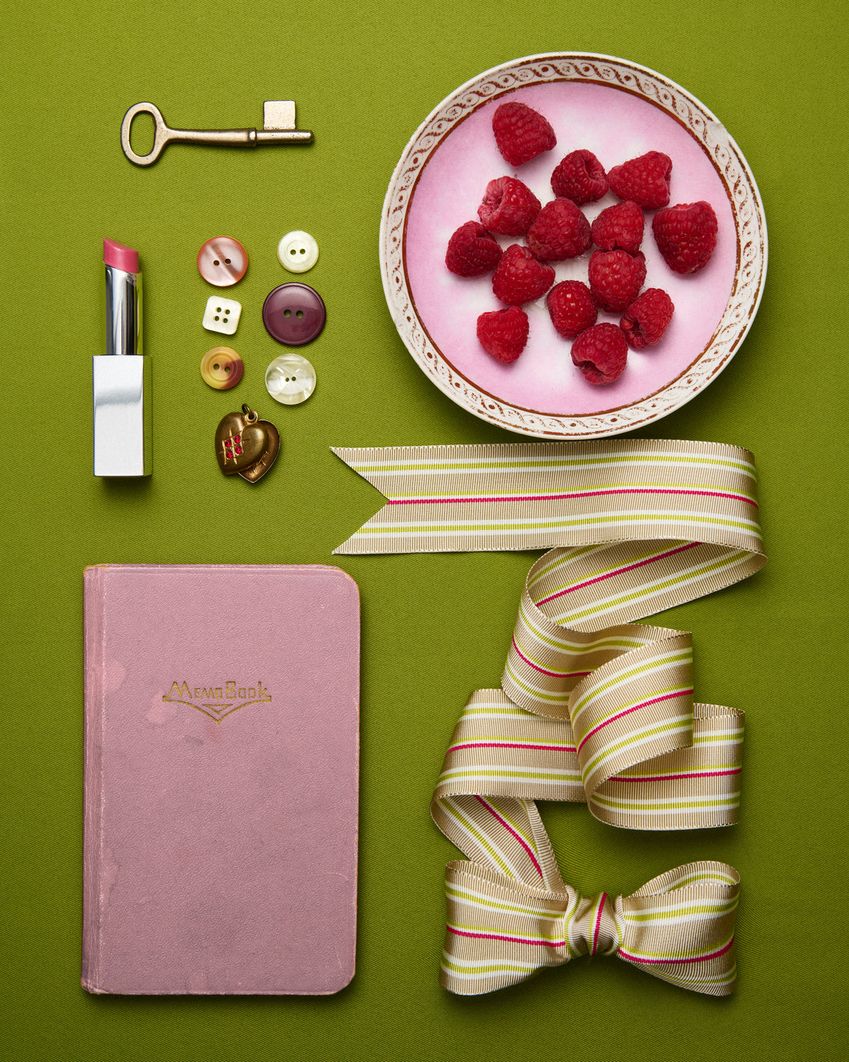 A styled flat-lay arrangement on a solid olive green background featuring a pink notebook with gold branding, a pink lipstick, a vintage-style key, buttons, a bowl of raspberries on a pink plate, and cream ribbon with red and green stripes.