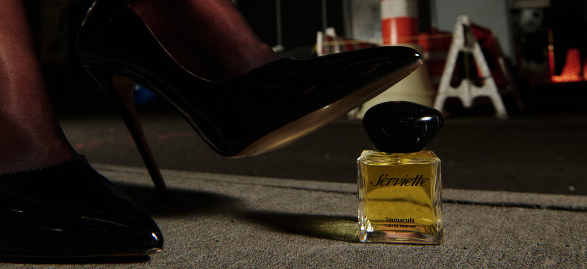 A close-up of a person's foot in a black patent leather high heel stepping on a bottle of Serviette perfume, which contains yellow liquid, set against a dark, industrial background.
