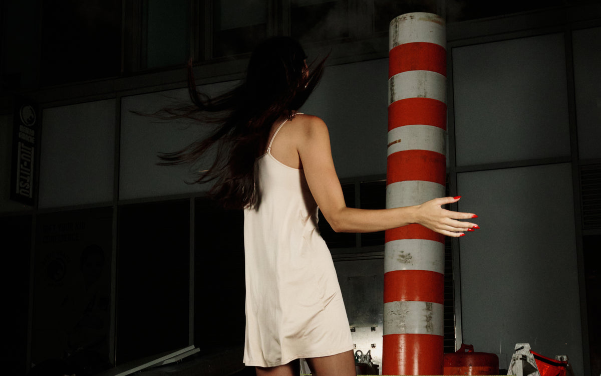 A woman in a sleeveless cream-colored dress stands with her back to the camera, reaching toward a red and white striped pole in a dark, nighttime urban setting.